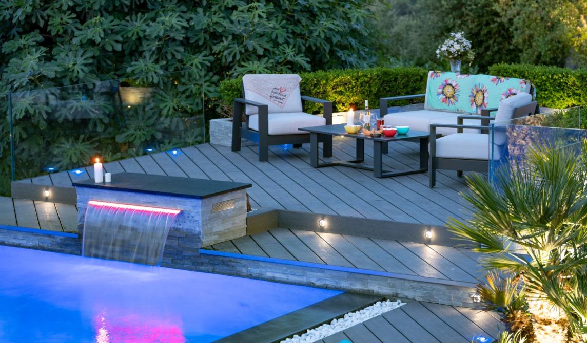 Terrasse en bois composite gris foncé avec salon de jardin moderne aux coussins blancs, bordure de piscine illuminée en bleu avec cascade, éclairage d'ambiance nocturne, végétation luxuriante et palmiers